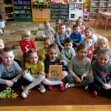 Dzieci siedzą na dywanie w sali przedszkolnej. Dziewczynka na pierwszym planie trzyma w rękach książkę z której bibliotekarka czytała opowiadania. 